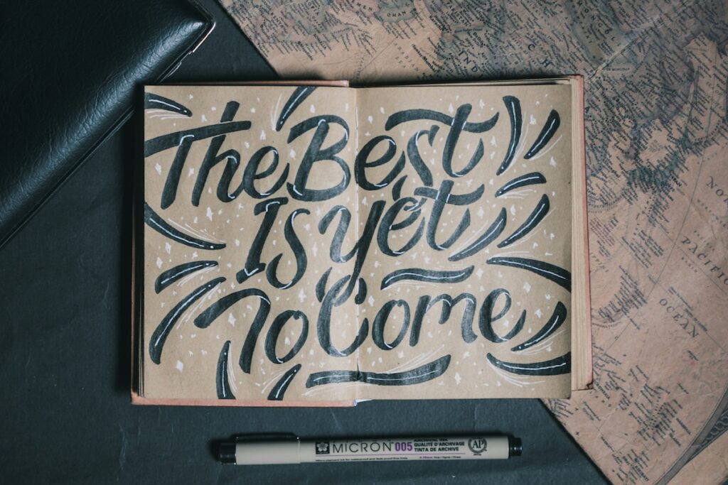 Open book with inspirational words drawn artistically stating "The Best Is Yet To Come" Surrounding the book is a map, black leather and a pen